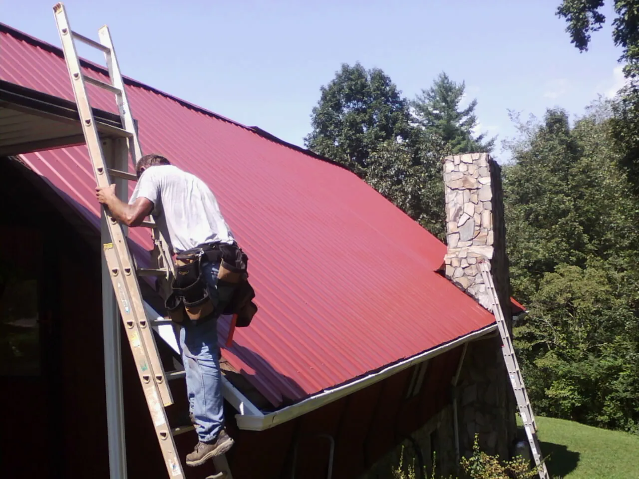Roofer on a ladder beside a red metal roof and stone chimney — Soffit & Fascia Repair by Roofing Brothers in Holden